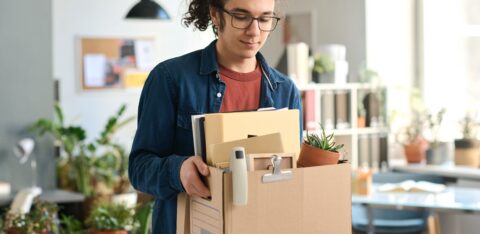 Young man in glasses carrying a box of personal items through an office.