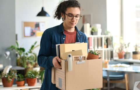 Young man in glasses carrying a box of personal items through an office.