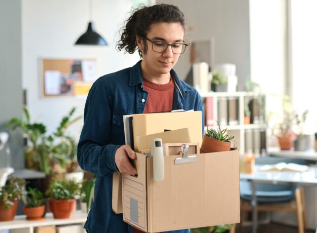 Young man in glasses carrying a box of personal items through an office.
