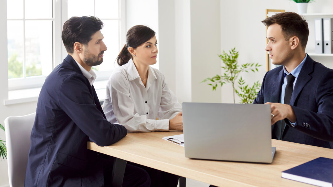 Couple sitting at a desk listening to a loan officer with a laptop in an office.