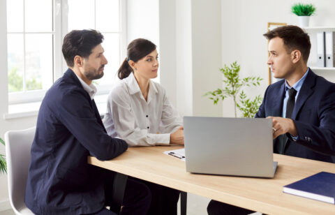 Couple sitting at a desk listening to a loan officer with a laptop in an office.